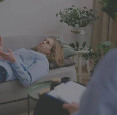 En person ligger på en soffa och förklarar något för en person i förgrunden.
