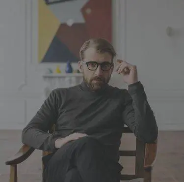 En man sitter i en stol med korsade ben framför en färgglad målning.