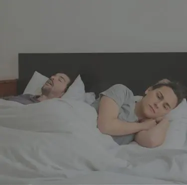 En man ligger i en säng och sover, med en vaken kvinna bredvid sig.