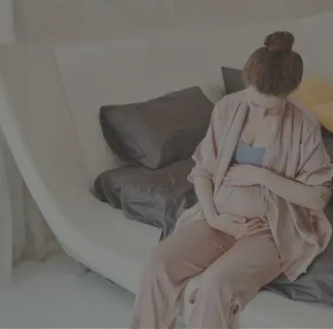 Gravid kvinna sitter på säng och håller sig för magen.